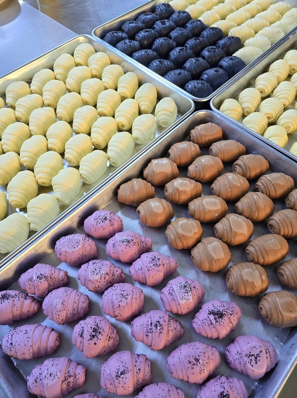Trays of hand-laminated croissant dough in charcoal, chocolate, taro and plain — proofing before the oven